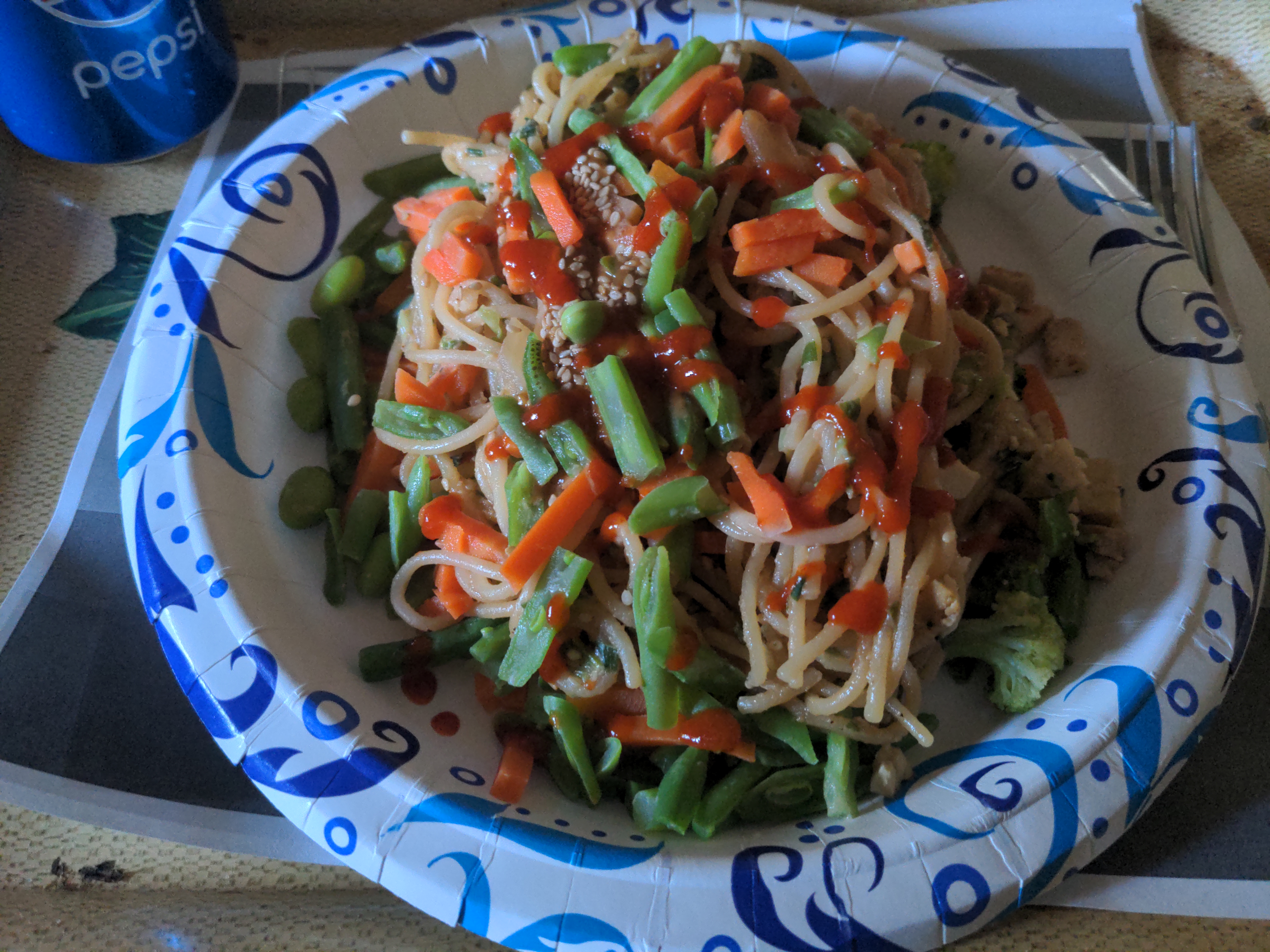 Amy's Chinese noodles and vegetables on a paper plate with sriracha sauce, and a can of Pepsi, on a TV tray